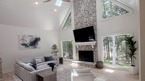 A spacious living room with a stone fireplace, a large TV, and a high ceiling with skylights.