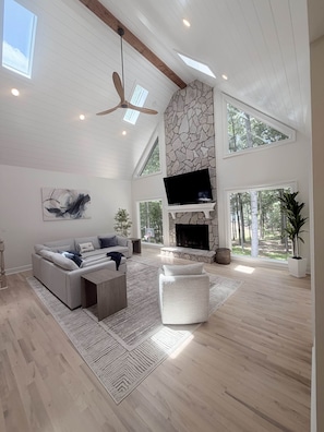 A spacious living room with a stone fireplace, a large TV, and a high ceiling with skylights.