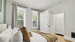 Une chambre à coucher avec un lit, deux fenêtres avec des rideaux, une porte blanche et un plancher en bois.
