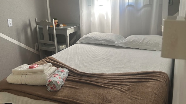 A single bed with a brown blanket, white pillows, and a floral pillow. There is a small table with a lamp and a decorative item. A window with sheer curtains allows natural light into the room. - Pousada e Hostel Flórida  (São Carlos)