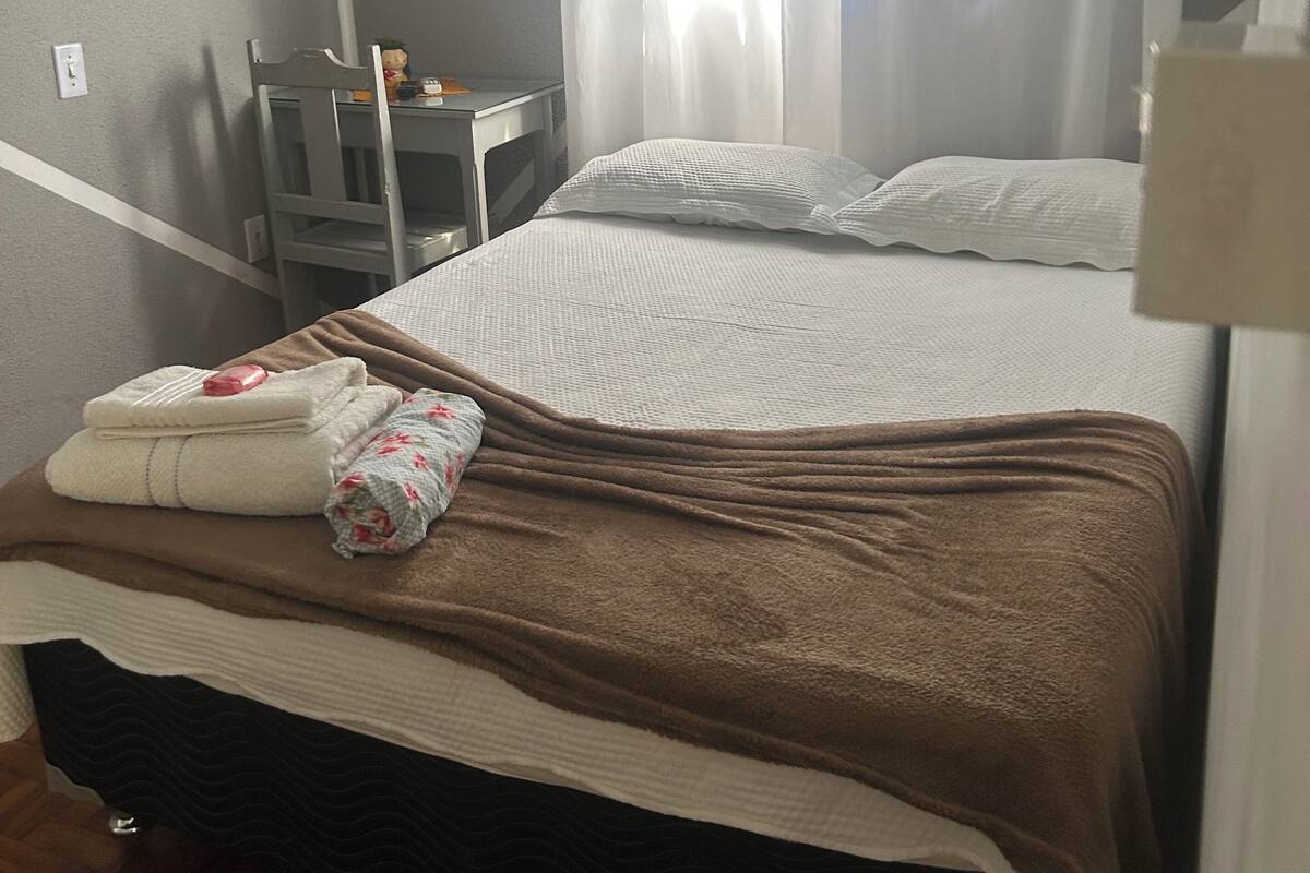 Een eenpersoonsbed met een bruine deken, witte kussens en een kussen met bloemenprint. Er staat een klein tafeltje met een lamp en een decoratief object. Een raam met dunne gordijnen laat natuurlijk licht de kamer binnen.