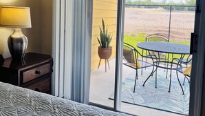 Habitación de hotel con cama, mesita de noche, lámpara y puerta corrediza de vidrio que da a un patio exterior con mesa y sillas.