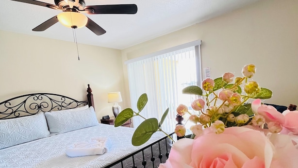 Un dormitorio con cama, ventilador de techo, ventana con persianas y un jarrón con flores.