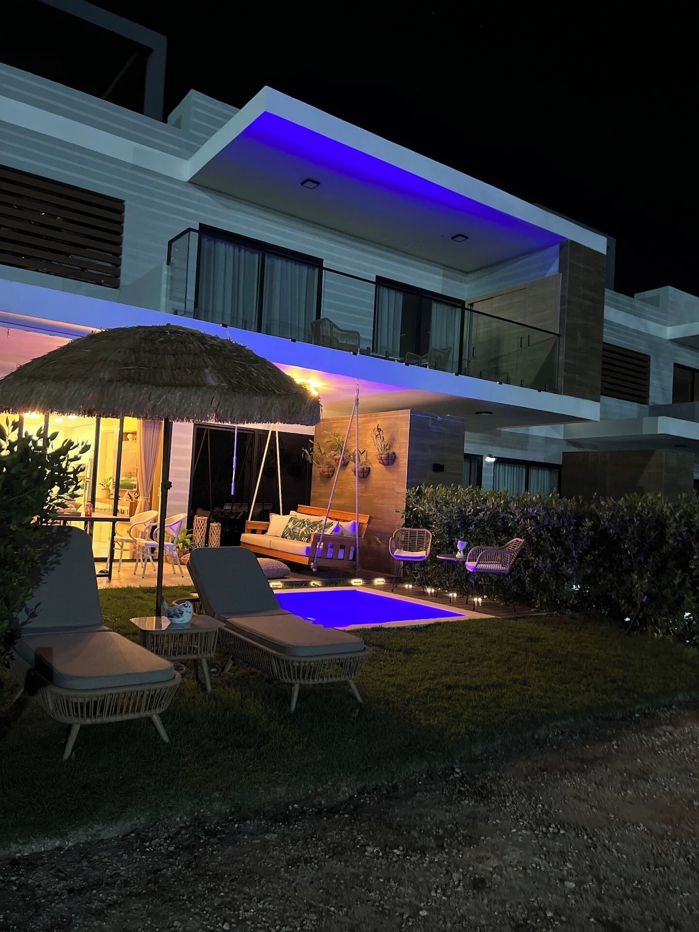 A modern two-story house with a swimming pool, outdoor seating, and a thatched umbrella.