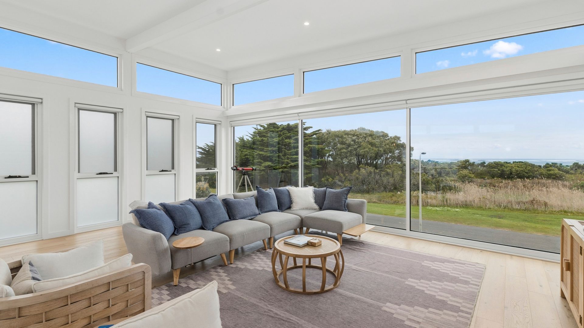 Uma sala de estar espaçosa com um sofá modular cinza, uma mesa de centro redonda de madeira e grandes janelas do chão ao teto com vista para o exterior.