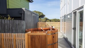 A wooden hot tub in a backyard with a wooden fence, a green house, and a white building with a sliding glass door. - Cedar Tides - Hot Tub & Waterfront (Apollo Bay)