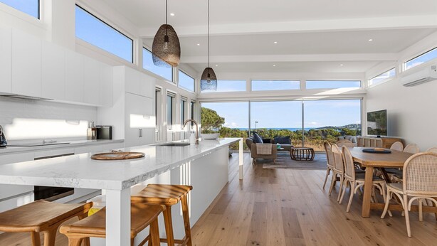 A modern kitchen with a large island, wooden flooring, and a dining area with wicker furniture. - Cedar Tides - Hot Tub & Waterfront (Apollo Bay)