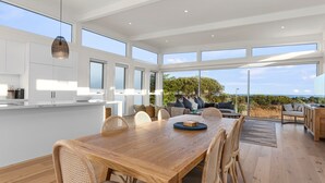 A spacious dining area with a wooden table and chairs, a kitchen with white cabinets, and large windows offering a view of the outdoors. - Cedar Tides - Hot Tub & Waterfront (Apollo Bay)