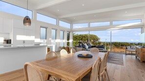 A spacious dining area with a wooden table and chairs, a kitchen with white cabinets, and large windows offering a view of the outdoors. - Cedar Tides - Hot Tub & Ocean Views (Apollo Bay)