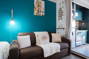 Ein Wohnzimmer mit einem braunen Sofa, einer Lampe, einem Gemälde und einem durch eine offene Tür einsehbaren Küchenbereich.