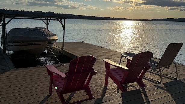 Una terraza de madera con dos sillas Adirondack y una mesa, con vista a una masa de agua tranquila donde un bote está amarrado cerca.