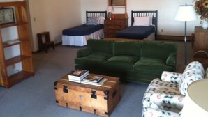 Una habitación con un sofá verde, un aparador de madera, una mesita con libros, dos camas con ropa de cama distinta, un espejo y una lámpara.