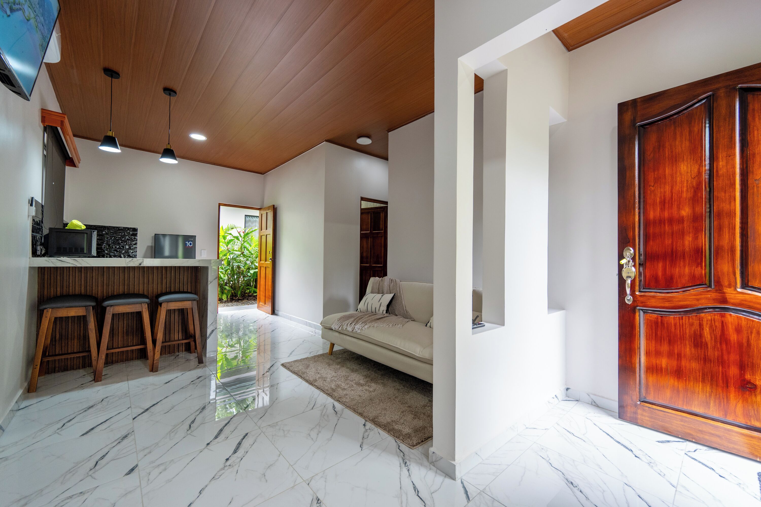 Un interior moderno con techo de madera, un sofá blanco, una puerta de madera y una zona de cocina con taburetes.
