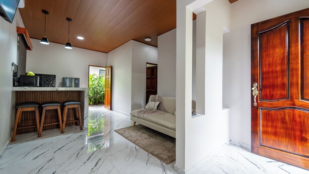 A modern interior with a wooden ceiling, a white sofa, a wooden door, and a kitchen area with stools. - River Collection - Villa Tranquil #3 sounds of the river (La Fortuna)