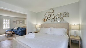 A bedroom with a large bed, a sofa, two bedside tables with lamps, a wall art piece, and a door leading to another room.
