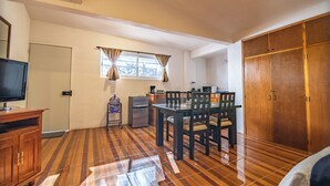Ein Zimmer mit Fernseher, Kühlschrank, Esstisch mit Stühlen, einem Fenster mit Vorhängen und einem Kleiderschrank.