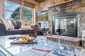 A cozy living room with a stone fireplace, a leather sofa, and a glass coffee table with fruit and a wine glass. - Moose Lodge: 4BR retreat with hot tub, fireplace, and game room. (Bethel)