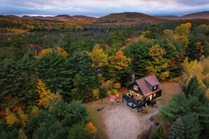 A cabin with a deck and outdoor seating, surrounded by dense forest and hills. - Moose Lodge: Hot Tub, Fireplace, Pet Friendly (Bethel)