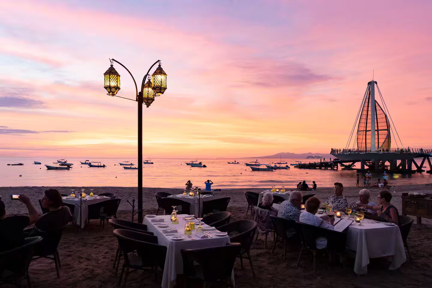 En restaurant ved stranden med borde klar til spisning, med udsigt over havet og en bro.
