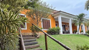 A house with a wooden staircase leading to a covered porch, surrounded by greenery and a well-maintained lawn. - Beautiful and comfortable house with security in a gated community near São Paulo. (Atibaia)