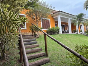 A house with a wooden staircase leading to a covered porch, surrounded by greenery and a well-maintained lawn.