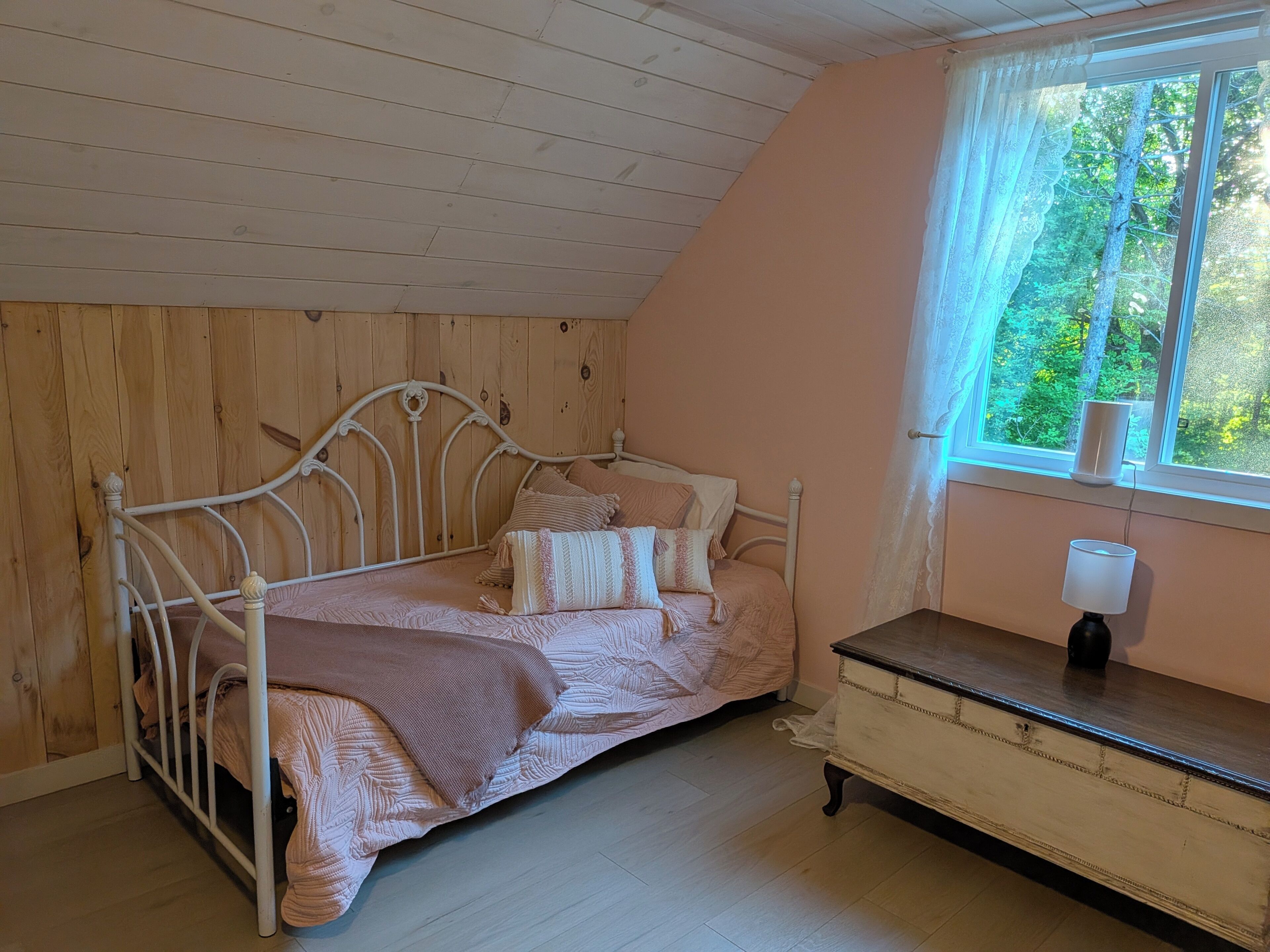 Un dormitorio con techo de madera, estructura metálica para la cama, mesita de noche con lámpara y ventana con vista a árboles.