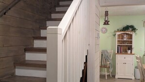 A wooden staircase with white railings, a wooden floor, a small cabinet with books, and a wall clock. - Cosy on-farm Cottage near Hardwood Lake. Fire pit, hiking trails, Beach nearby (Palmer Rapids)