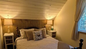 A bedroom with a wooden headboard, a bed with a white quilt, two bedside tables with lamps, and a window with curtains. - Cosy on-farm Cottage near Hardwood Lake. Fire pit, hiking trails, Beach nearby (Palmer Rapids)