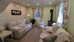 A cozy living room with a sofa, a chair, a table, a lamp, a plant, a fireplace, and a window with curtains. - Cosy on-farm Cottage near Hardwood Lake. Fire pit, hiking trails, Beach nearby (Palmer Rapids)