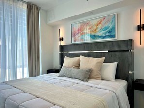 A modern bedroom with a large bed, a headboard with vertical lines, a painting above the bed, and a nightstand with a lamp. - X Lounge IX-Signature Skyline Residence (Miami)
