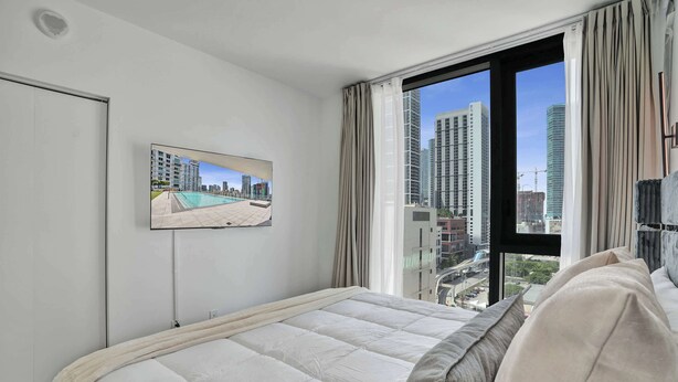 Quarto de hotel com uma cama, televisão na parede e uma janela grande com cortinas.