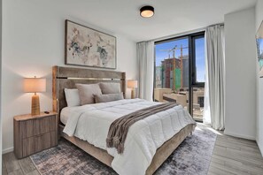 A hotel room with a large bed, a nightstand, a lamp, a painting, and a view of a construction site through the window. - X Lounge IX Signature | 3 BR/3 Ensuite Residence (Miami)