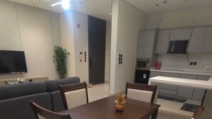 A modern living room with a dark wooden dining table, four chairs, a grey sofa, a flat-screen TV, and a kitchen area with grey cabinets and a microwave. - OrbitNet - A05 (Madina)