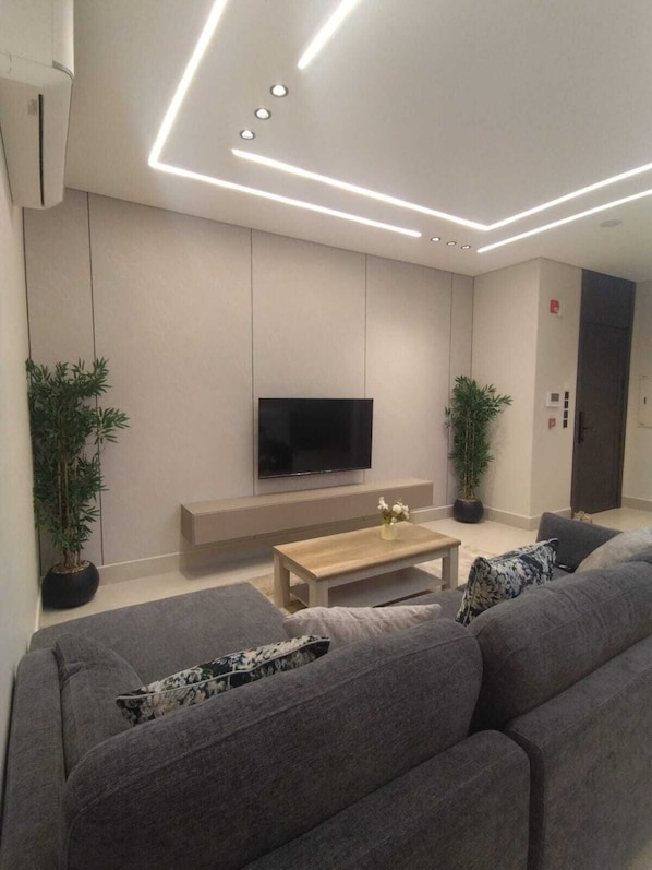 A modern living room with a flat-screen TV, grey sofas, a wooden coffee table, and two potted plants.