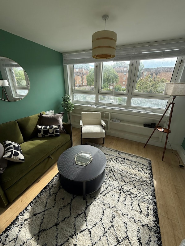 A living room with a green sofa, a round coffee table, a white armchair, a floor lamp, and a large window with a view of buildings.