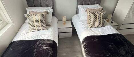 A hotel room with two beds, each with a grey headboard and white bedding. There are two bedside tables with lamps and a small box on each.