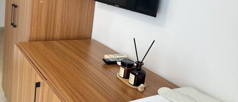 Un mueble de madera con un televisor montado en la pared encima, una mesita con una botella y una vela, y una almohada blanca sobre el mueble.