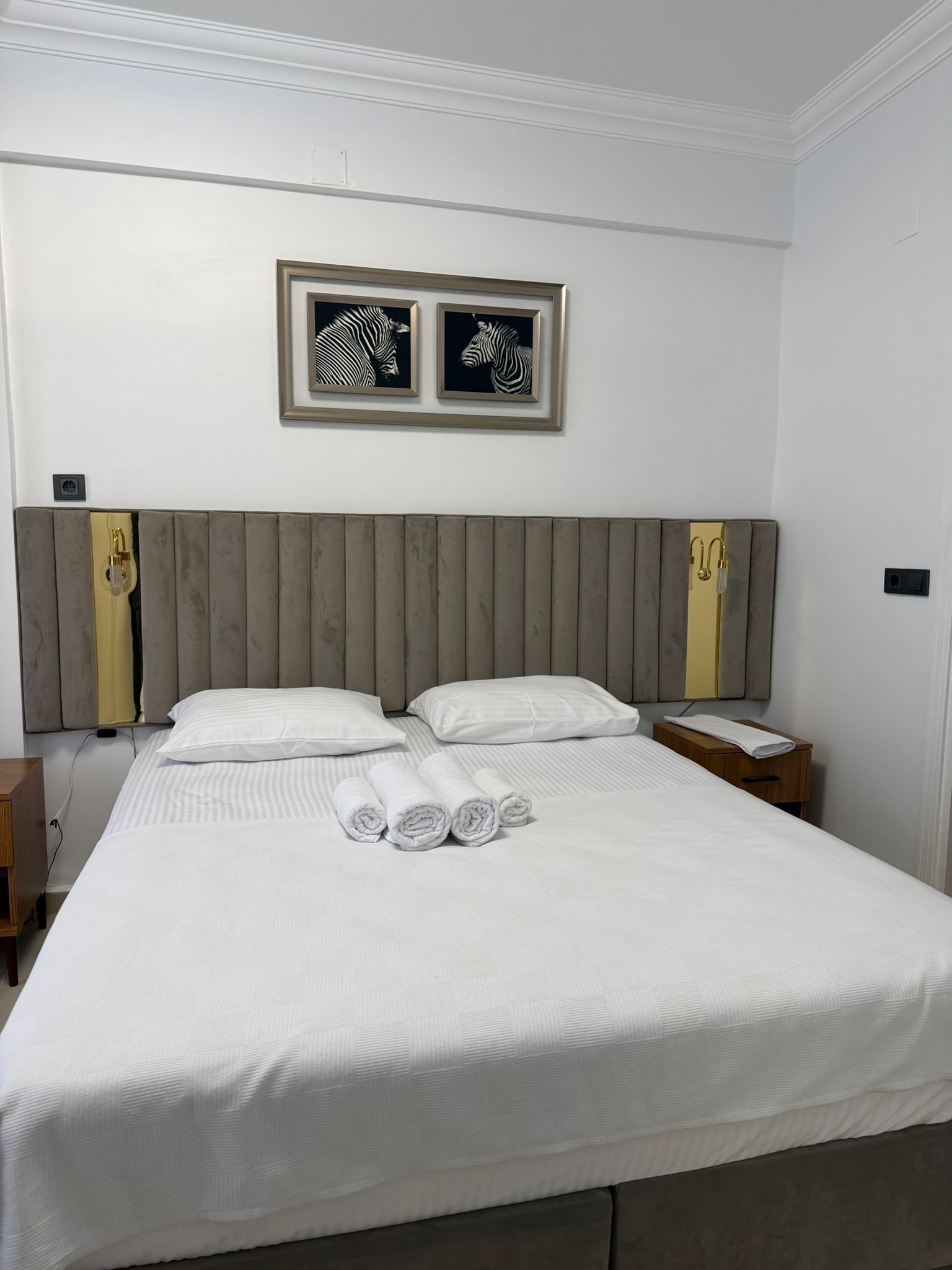 A hotel room with a large bed, two pillows, a headboard, a nightstand, a bedside table, and two framed zebra pictures on the wall.