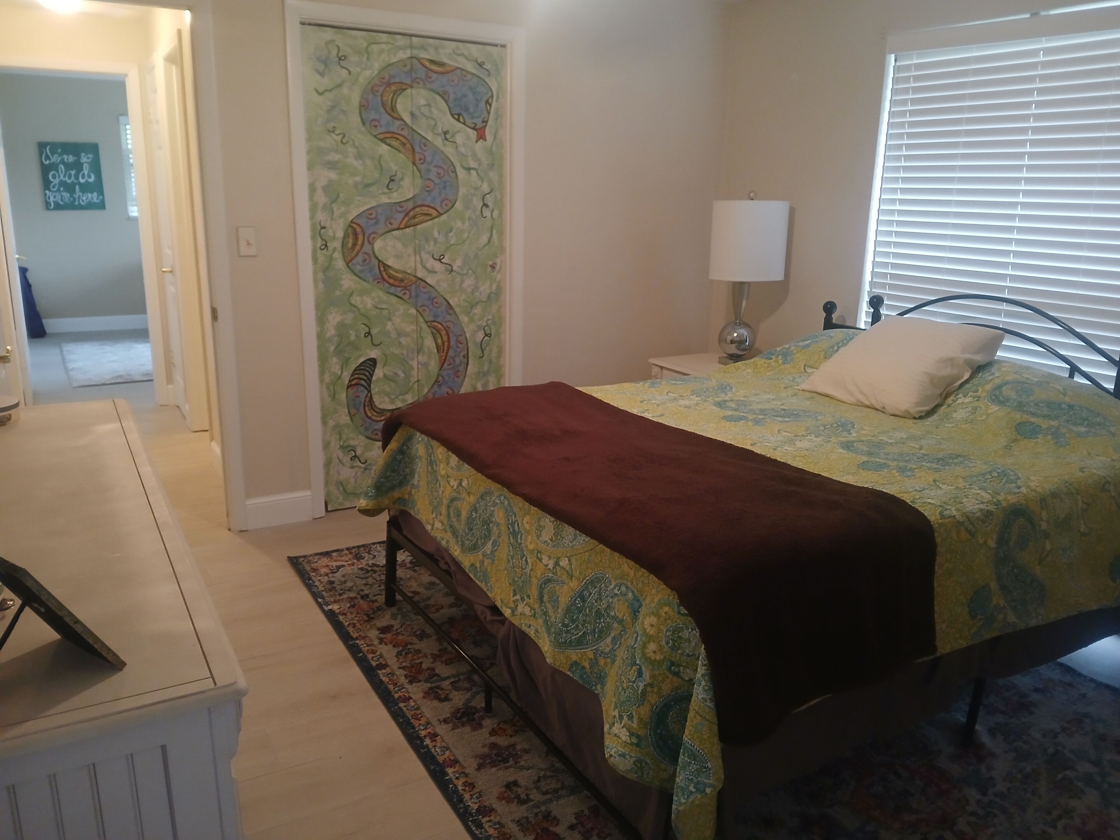 Quarto com cama, mesa de cabeceira, abajur, porta com design de cobra e tapete.