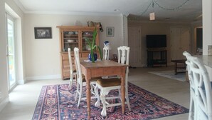 Een eetgedeelte met een houten tafel, witte stoelen, een kamerkleed met patroon, een kast en een televisie op de achtergrond.