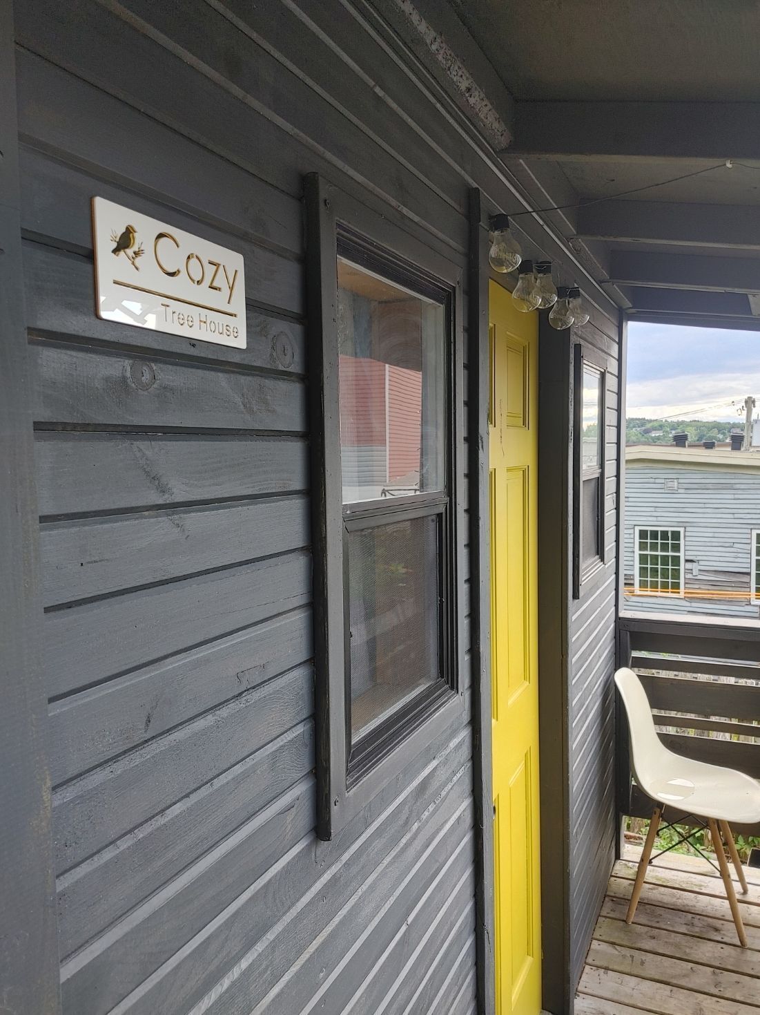 A building with a 'COZY Tree House' sign, a window with a mesh screen, a yellow door, and a white chair on a wooden deck.