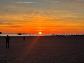 Ein Strand mit einem Steg, der sich ins Wasser erstreckt, einem Sonnenuntergang im Hintergrund und Silhouetten von Personen im Sand.