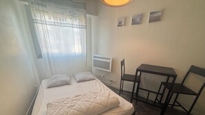 A single bed with a white duvet, a small table, two chairs, a TV, and a window with a curtain.