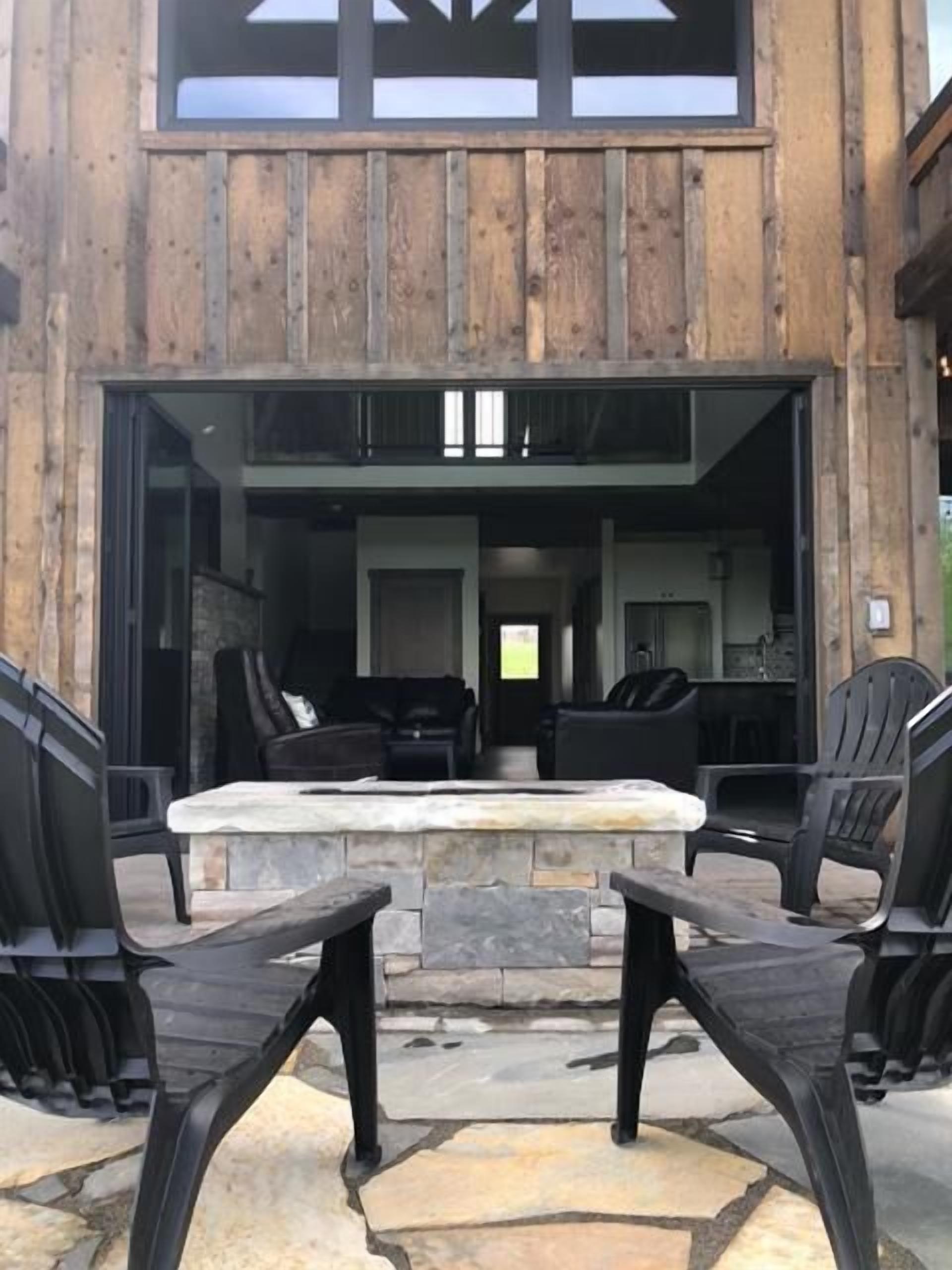 An outdoor seating area with stone tables and chairs, a stone fireplace, and a view into a building with a wooden facade.