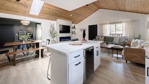 A modern kitchen with a central island, a dining area with benches, a living room with a sofa, and a TV mounted on the wall. - Maverick`s Cabin - HotTub, GameRm, Firepit, fenced (Big Bear)
