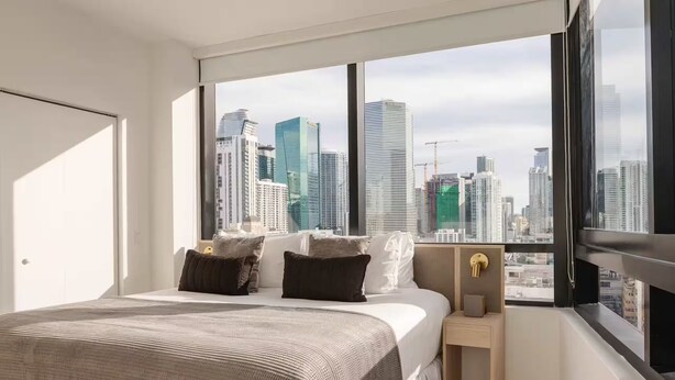 Camera moderna con un letto grande, un comodino e vista su un orizzonte cittadino attraverso ampie finestre.
