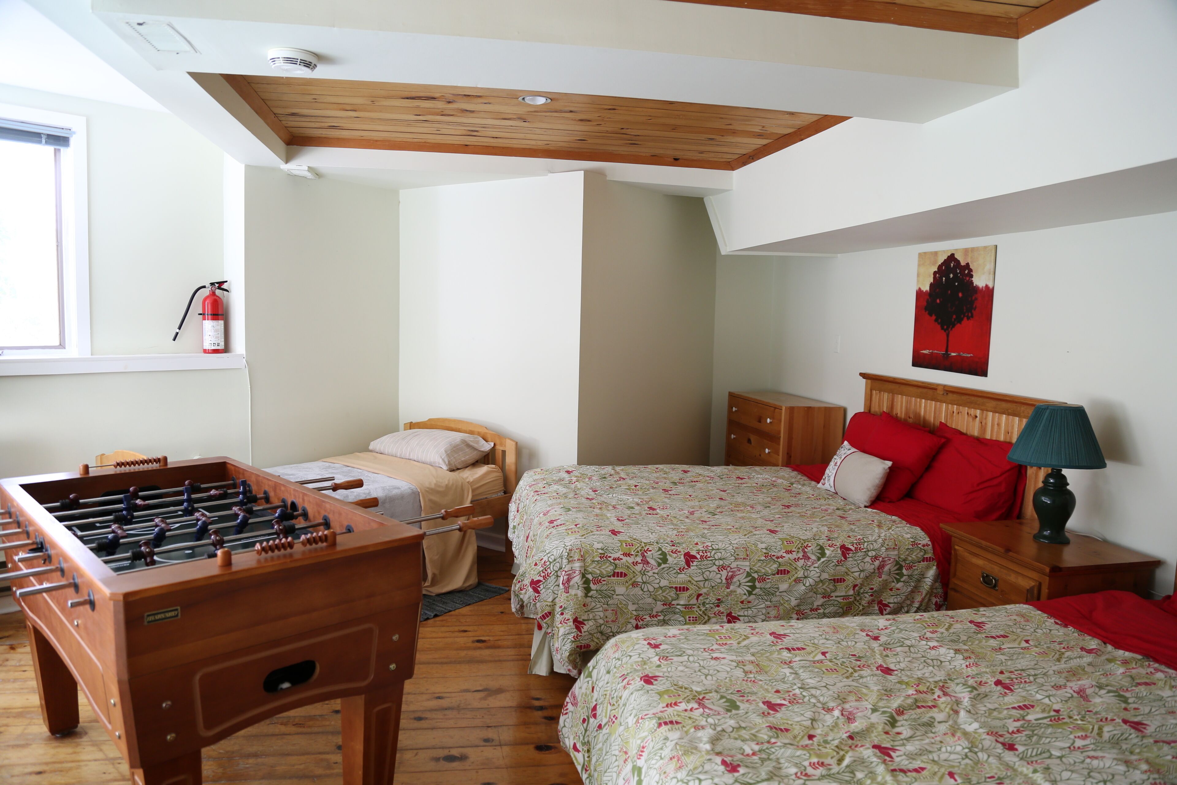 A room with a foosball table, two beds with floral bedding, a wooden ceiling, a fire extinguisher, and a painting on the wall.