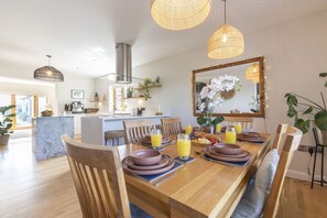 A dining area with a wooden table set for four, surrounded by chairs, with a kitchen in the background. - Beautiful 4 Bed 3 Bath Cotswold Home with Hot Tub (Cheltenham)