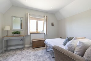 A bedroom with a bed, bedside table, lamp, window, and a small wooden bench. - Beautiful 4 Bed 3 Bath Cotswold Home with Hot Tub (Cheltenham)