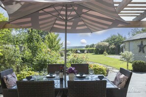 Uma área de refeições ao ar livre com móveis de vime, uma mesa posta para dois e um grande guarda-sol oferecendo sombra.